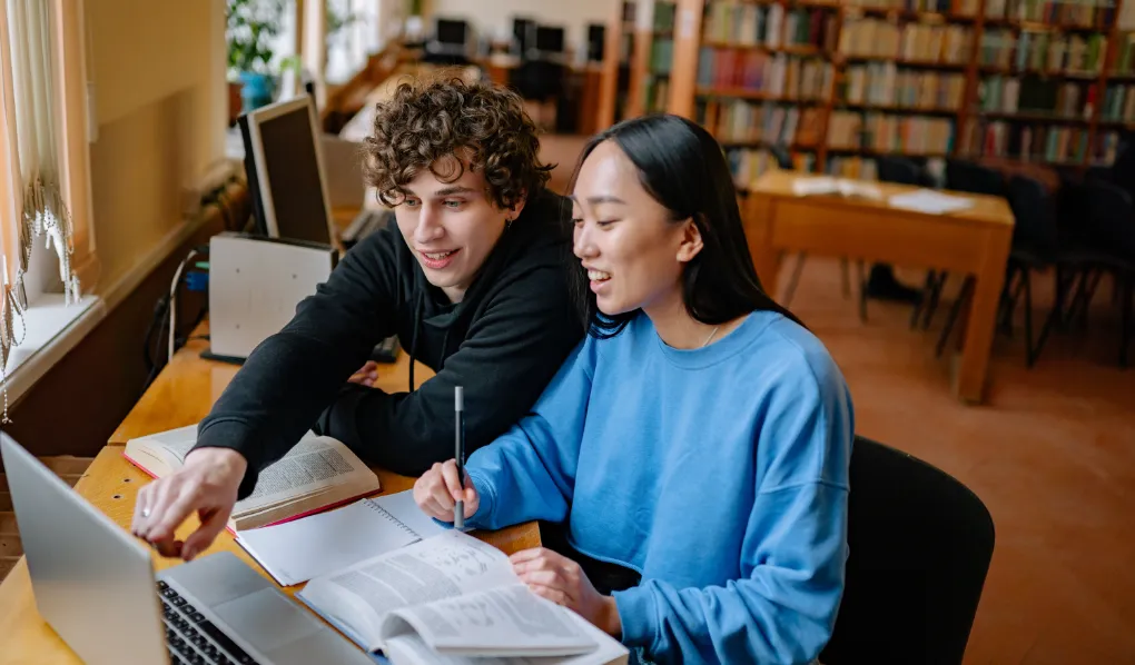 Two online students studying in library and smiling at laptop