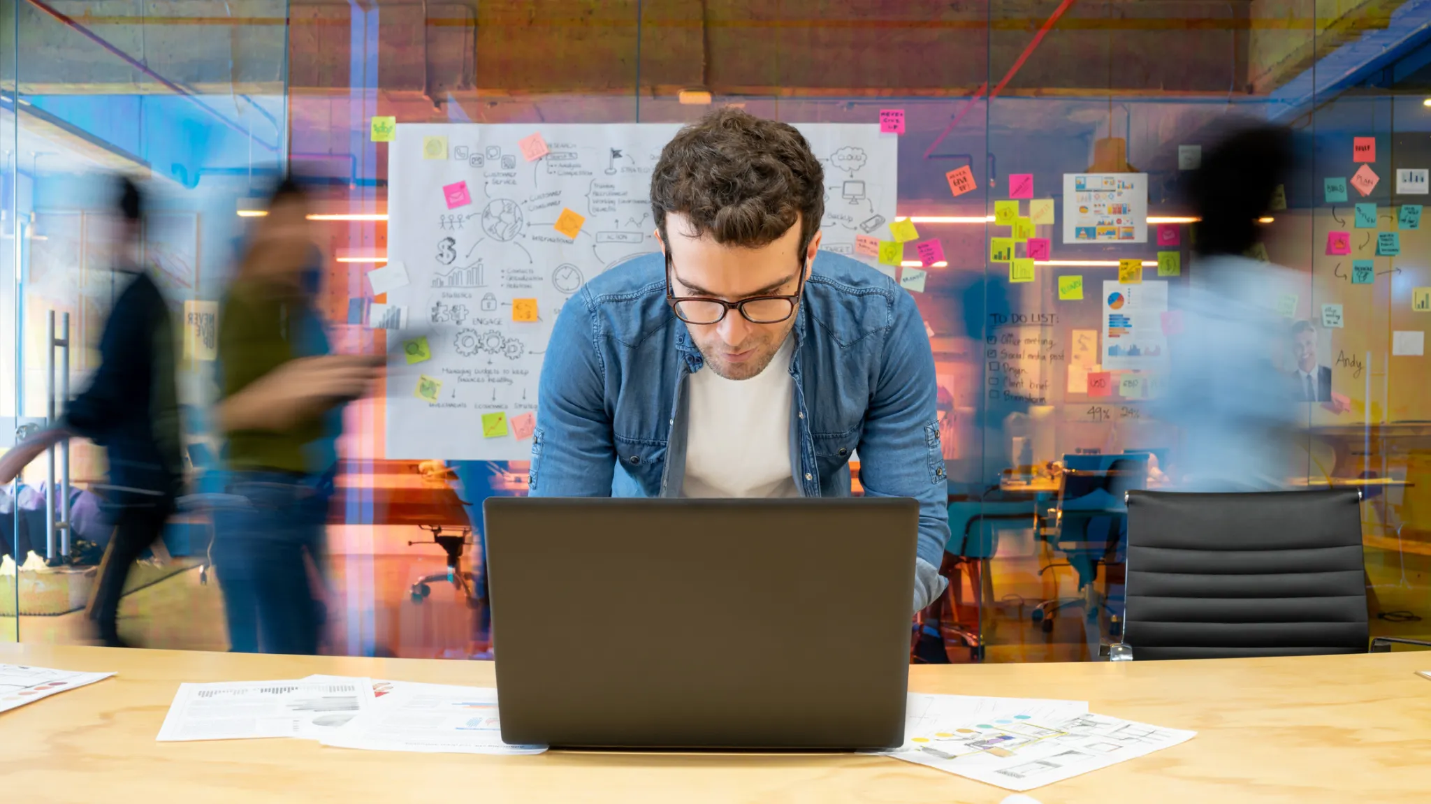 Young man with glasses working on his laptop with a strategy board behind him